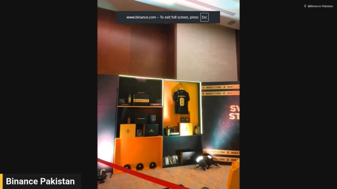 Photo from a Binance Pakistan event showing a display with a framed jersey, branded merchandise boxes, and a 'www.binance.com' banner at the top of a stage setup.