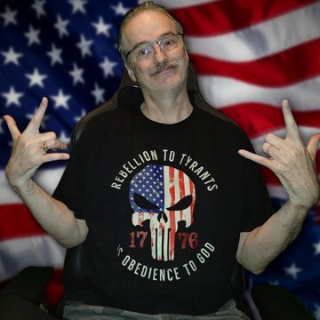 A man wearing glasses and a black t-shirt reading 'REBELLION TO TYRANTS 1776 OBEDIENCE TO GOD' with a skull and American flag design, posing in front of an American flag backdrop.