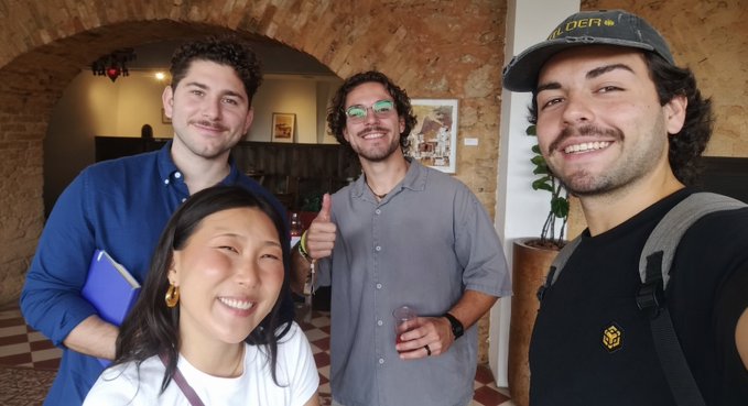 A selfie of four smiling people posing together in what appears to be a brick archway interior, with one person wearing a blue shirt, another in a gray button-up, a woman in white in front, and a man in a black shirt with a cap on the right.