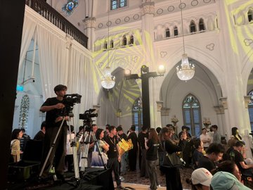 An interior view of a church or cathedral during an event, with a cameraman filming and people gathered near a large illuminated cross and chandelier.