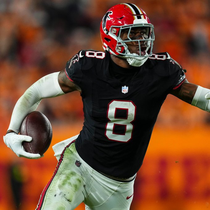 An American football player wearing an Atlanta Falcons black jersey with number 8 carries the ball down the field against an orange stadium background.