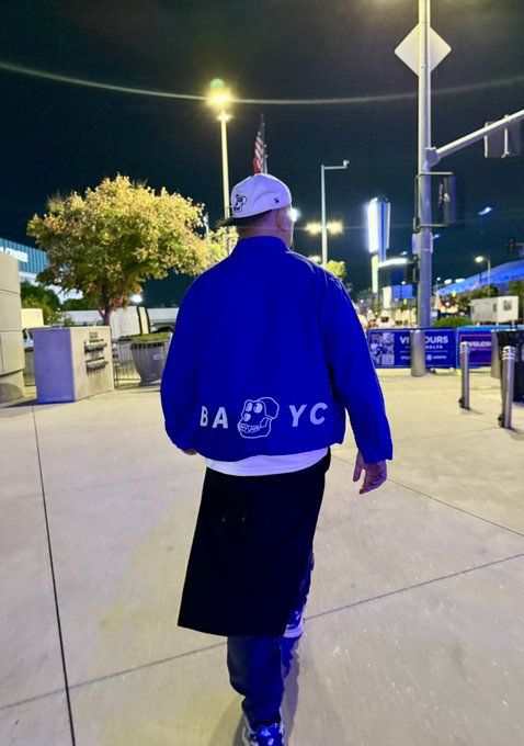 A person wearing a white cap and a blue "B.A.Y.C." bomber jacket with a cartoon face on the back walks through an outdoor area at night near what appears to be a stadium entrance.