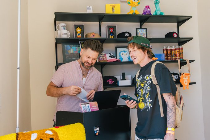 Two men interact at what appears to be a retail counter, with shelves behind them displaying merchandise including "Don't F*ck With Cats" and "Gentleman's Club" hats.