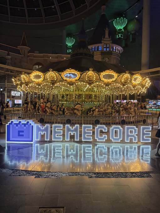 An illuminated 'MEMECORE WELCOME' sign in pixel font stands in front of an ornate, lit-up carousel at night.