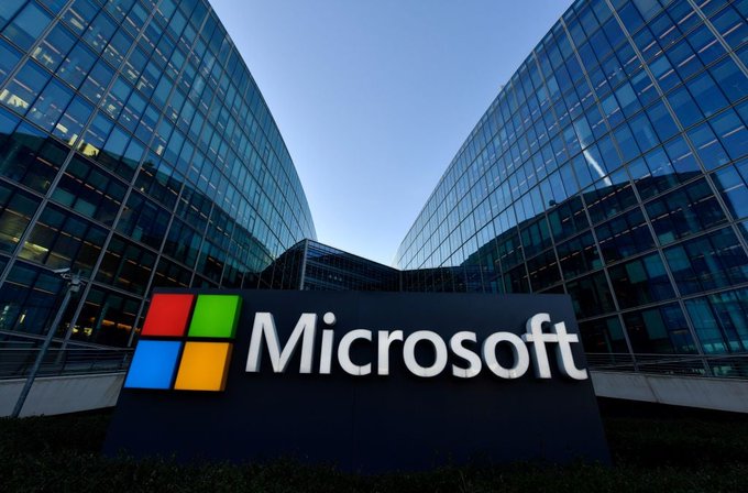 The Microsoft corporate logo stands prominently in front of the company's modern glass office buildings, accompanying news that Microsoft ($MSFT) is laying off approximately 4,800 workers—roughly 2% of its workforce—with HR noting the cuts are not being replaced by AI.