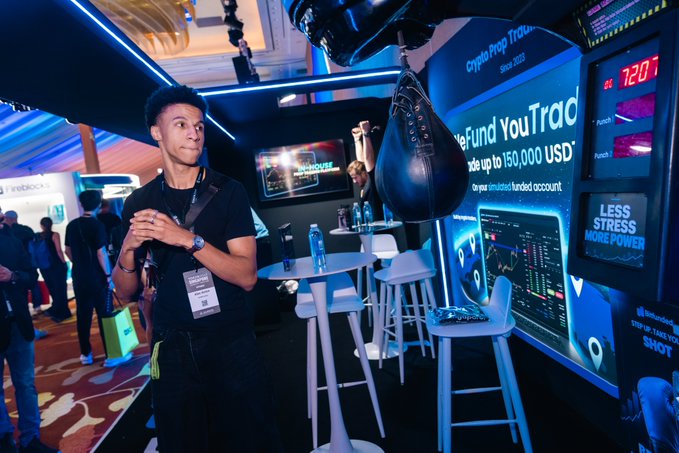A young man at a brightly lit expo booth stands near a hanging speed bag, with signage reading 'Crypto Prop Trading,' 'We Fund You Trade,' 'Up to 150,000 USD,' and 'LESS STRESS MORE POWER.'
