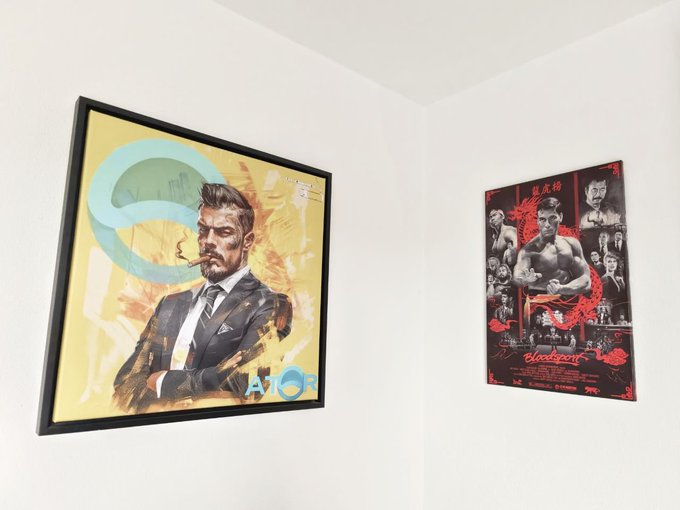 Two framed posters hang on a white wall, one featuring a suited man with a cigar and the text 'AMON', the other a Bruce Lee 'Bloodsport' style poster with red Chinese characters.