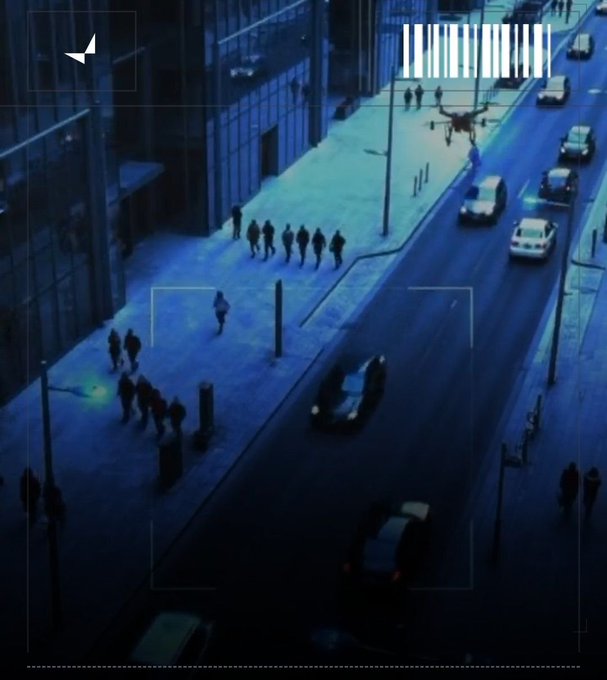 An aerial, blue-tinted surveillance-style view of a city street with pedestrians walking on the sidewalk and cars driving by, overlaid with targeting reticle graphics.