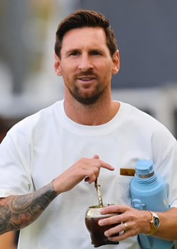 A bearded man wearing a white t-shirt and a watch holds a mate gourd with a blue thermos, with tattoos visible on his forearm. The image relates to the post about Lionel Messi receiving a farewell match with Argentina, capturing him in a casual moment likely during his downtime with teammates.