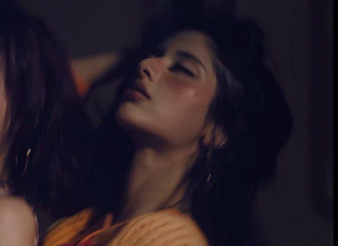 A young woman with long dark hair and closed eyes leans back in a dimly lit, moody setting, wearing large hoop earrings and an orange-toned garment. The intimate, cinematic shot reflects the emotional or evocative tone often associated with musical artists tagged in the post, "ARIA" and "XIN."