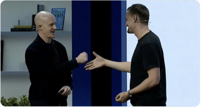 Two men shaking hands in what appears to be a studio setting, with a bookshelf visible in the background.