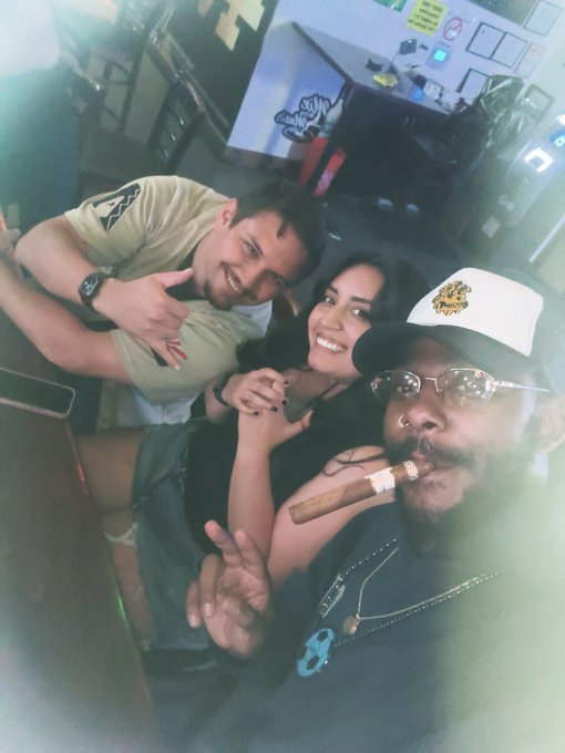 Three people pose for a selfie in a dimly lit venue, with a man in the foreground smoking a large cigar and wearing a black and white cap, gold glasses, and a pendant necklace.