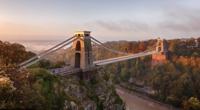 The Clifton Suspension Bridge in Bristol, England, spans a gorge surrounded by autumn-colored trees at sunrise, with the River Avon visible below.