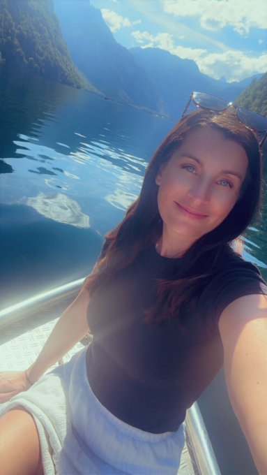 A selfie of a smiling woman with sunglasses on her head, sitting on a boat on a bright blue lake surrounded by green mountains under a partly cloudy sky.