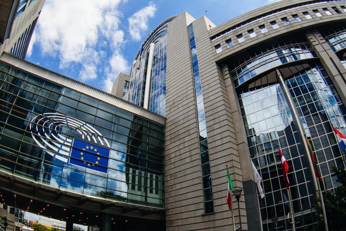 The image shows the European Parliament building in Brussels, featuring its distinctive curved glass facade with the EU flag and the Parliament's emblem prominently displayed. This relates to the post as it depicts the EU institution responsible for reviewing and potentially refining the MiCA (Markets in Crypto-Assets) regulatory framework governing stablecoins, tokenization, and digital assets in Europe.