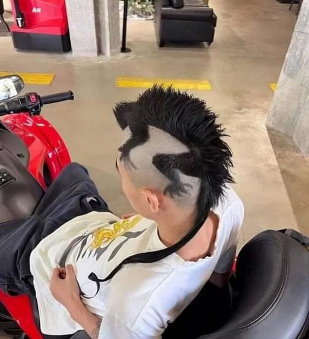 A person sits on a red motorcycle displaying an elaborate and edgy haircut featuring a spiky mohawk with shaved geometric patterns on the sides.