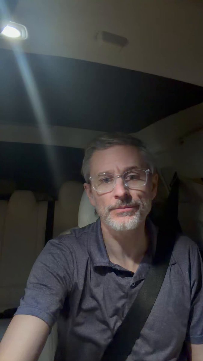 The image features a man wearing glasses, sitting inside a car with his head resting against the seatbelt. He appears to be looking directly into the camera, possibly posing for a photo or video. The background of the image shows a white wall, which contrasts with the dark ceiling and adds depth to the scene. There is also an airplane logo visible in the background, suggesting that they might be on a trip or vacation.