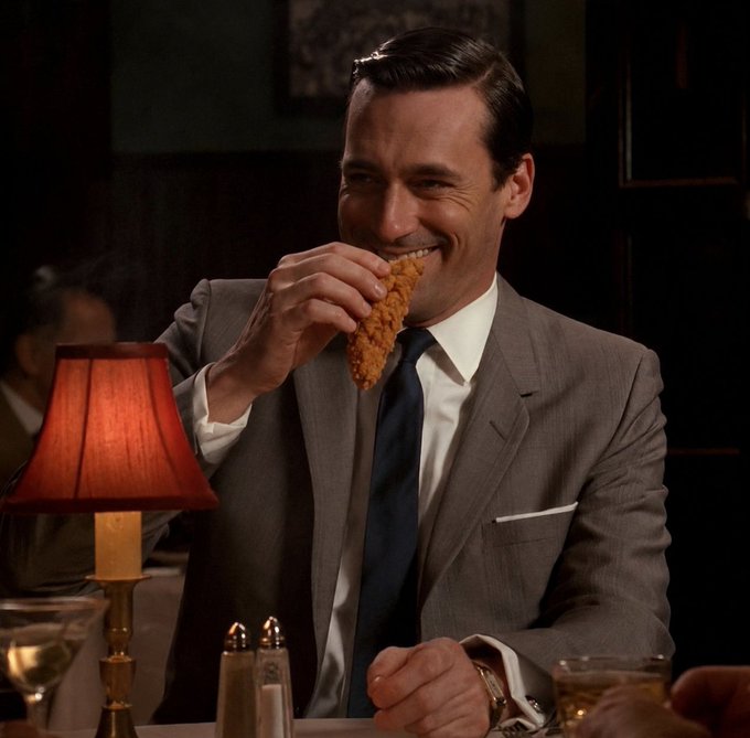 A man in a grey suit, white shirt, and dark tie is smiling while eating fried chicken at a dimly lit restaurant table.