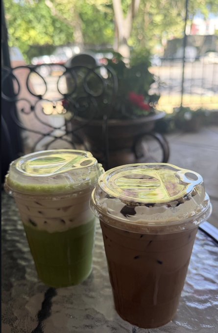 Two iced beverages in clear plastic cups with domed lids—one a green matcha latte and the other a brown iced coffee—sit on a textured table with a wrought-iron railing and street view in the background.
