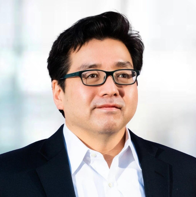 A professional headshot of an Asian man wearing black-rimmed glasses and a black suit jacket over a white collared shirt, smiling slightly against a blurred light background.