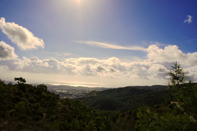 A scenic landscape photograph showing a vast view of forested hills and a distant coastline under a bright, cloudy sky with the sun shining overhead.