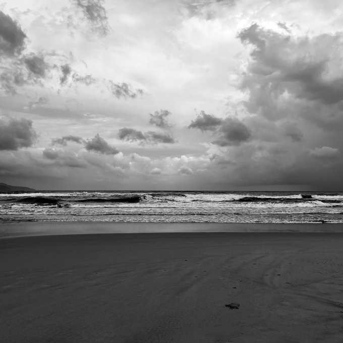 A moody black-and-white photograph of a deserted beach with waves rolling onto the sand under a heavy, cloud-filled sky.