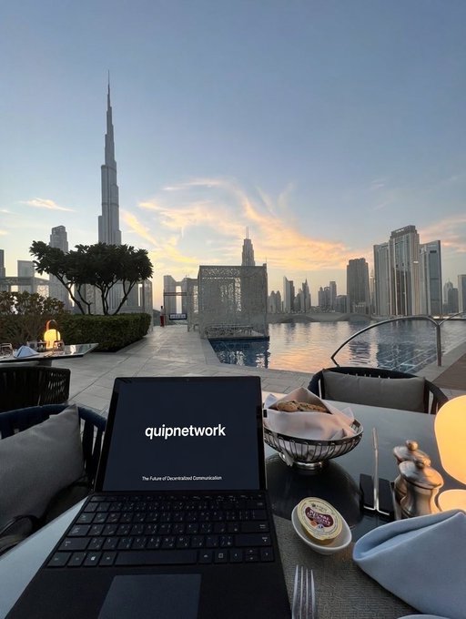 The image shows an open laptop computer sitting on top of a table with food items such as bread, butter, cheese, and other condiments. The scene takes place at a restaurant or dining area overlooking a city skyline, including the Burj Khalifa in Dubai. The setting appears to be a quiet evening where someone is working or studying while enjoying their meal.