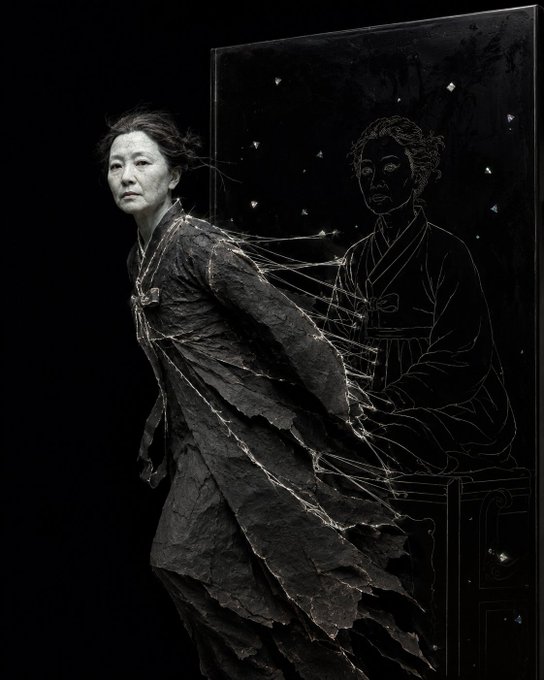 An elderly woman in tattered dark clothing stands beside an ornate black mirror or panel that reflects a drawn portrait of herself in a kimono.