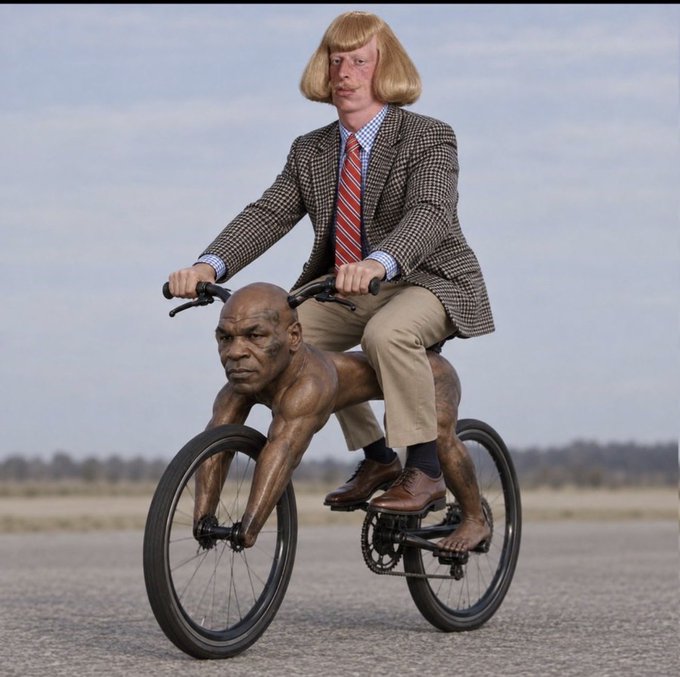 A surreal image showing a man in a checkered blazer and blonde wig riding a bicycle whose frame is shaped like Mike Tyson's muscular body, posed on a road under a blue sky.