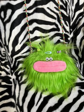 A fuzzy green monster plush purse/bag with a pink mouth, small eyes, and a green hair clip, hanging on a braided strap against a zebra-print background.