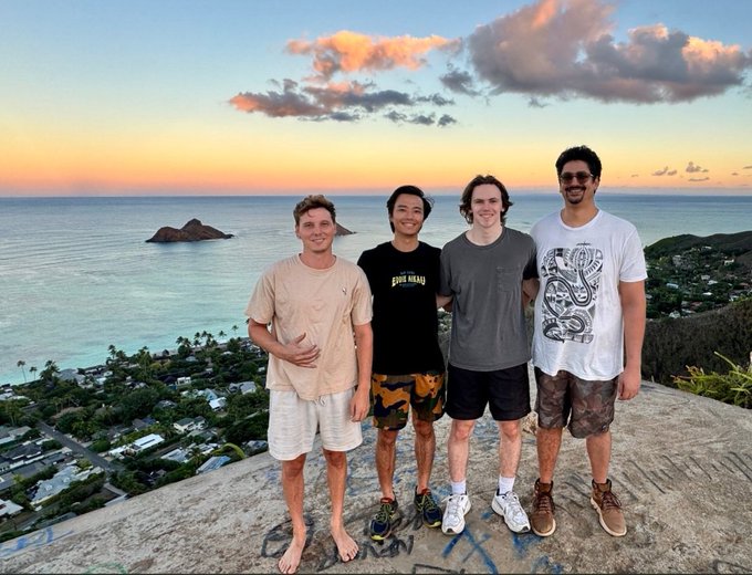 Four young men posing together on a rocky cliff at sunset, with a beautiful ocean view and small island visible in the background.