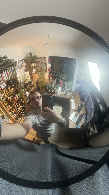 A man wearing glasses takes a selfie in a round mirror, capturing a reflection of a room filled with bookshelves, plants, and a desk cluttered with items.