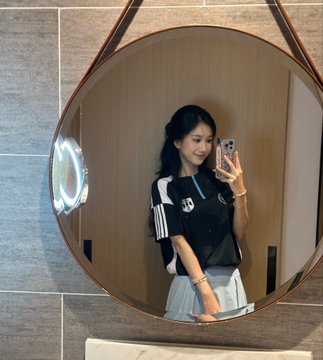 A young woman takes a mirror selfie in a round mirror, wearing a black and white striped jersey paired with a light-colored skirt, holding up her phone to capture the moment. Her sporty outfit complements the post's theme of the "#Bitget Midnight Football Party 2.0," suggesting she's dressed up to get hyped for the exciting match ahead.