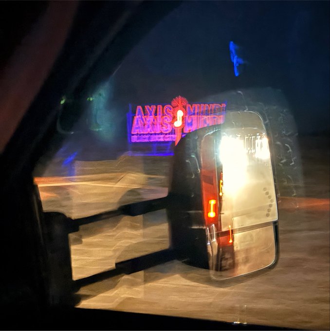 Nighttime photo taken from inside a vehicle, showing a glowing neon sign reading 'AXIS MUNDI' visible through the windshield alongside the reflection of headlights and side mirror.