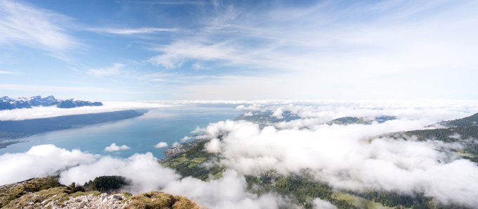 A scenic panoramic view of a large blue lake or fjord surrounded by mountains, partially obscured by white clouds and mist rolling over the landscape under a blue sky with wispy clouds.