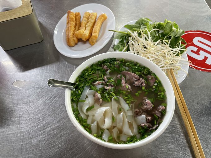 A bowl of Vietnamese pho with wide rice noodles, beef, and chopped green onions, served with a side of fried dough sticks (quẩy) and a plate of fresh herbs and bean sprouts on a metal table.