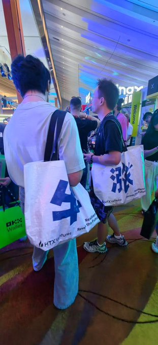 Two attendees at TOKEN2049 carry HTX (HTXVentures) branded tote bags featuring large blue Chinese characters "牛" (bull) and "涨" (rise) on a white background, lining up at the booth to collect the popular promotional merchandise.