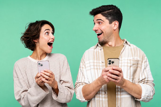 A young man and woman stand against a green background, both holding smartphones and looking at each other with expressions of surprise and excitement.