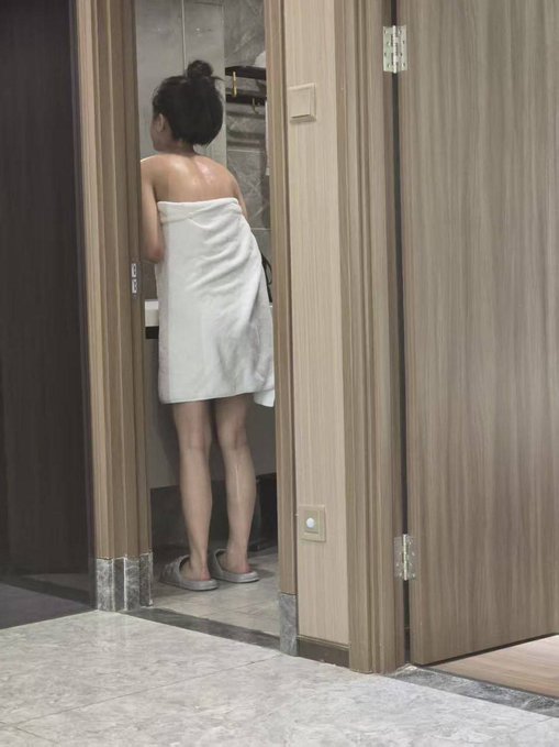 A woman wrapped in a white towel standing in a bathroom doorway, wearing slippers, with her hair in a bun.