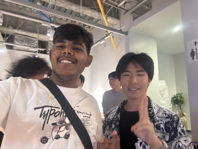 A selfie featuring two young men smiling indoors, with the person on the right making a peace sign, taken in what appears to be a modern cafe or event space.