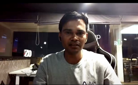 A webcam photo of a young man in a gray t-shirt sitting in front of a computer setup with monitors displaying '70' in the background.