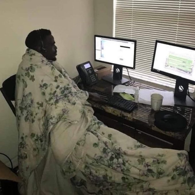 A man sitting at a desk with dual monitors and a phone, wrapped in a floral blanket and wearing a headset with a microphone.