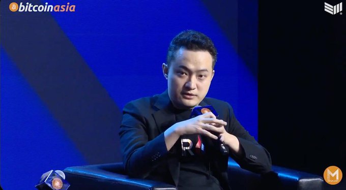 A man in a black suit speaks into a microphone while seated on a panel at a 'bitcoin asia' event, with an orange Bitcoin logo visible on the microphone and a blue backdrop behind him.