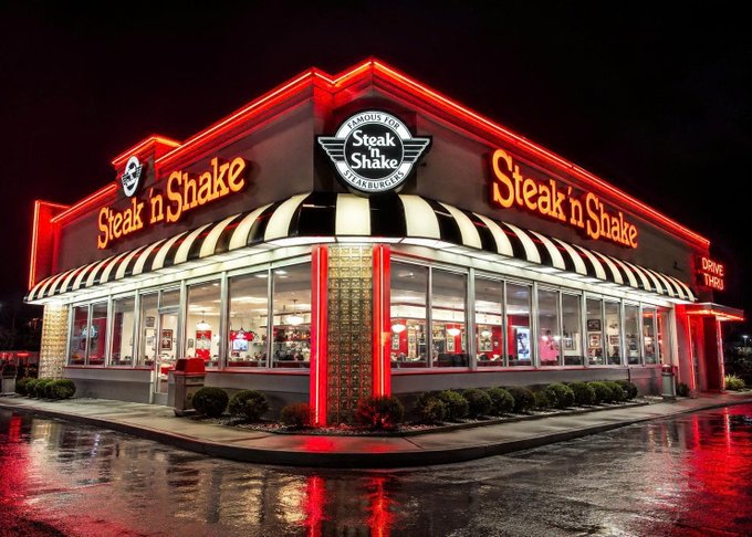 A Steak 'n Shake restaurant exterior at night features bright red neon lighting and black-and-white striped awnings.
