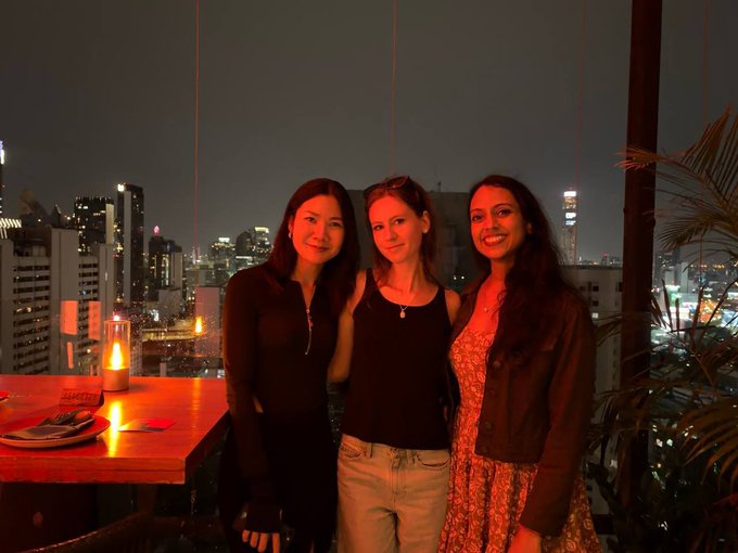 Three women posing together at an outdoor rooftop bar at night with a city skyline illuminated in the background.