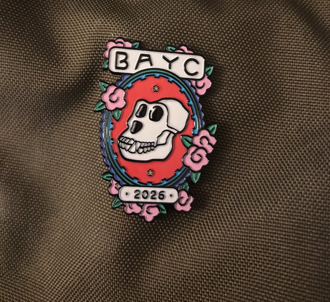 A decorative enamel pin featuring a skull design with "BAYC" at the top and "2026" at the bottom, surrounded by pink roses and a red oval frame, displayed on a textured olive-green fabric. The "BAYC" likely refers to Bored Ape Yacht Club, a popular NFT collection, with the 2026 possibly indicating an anniversary year, tying into the post's casual mention of "this one" as another collectible in a series.