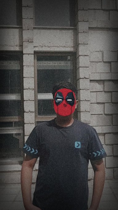 A masked figure wearing a red Deadpool-style mask poses in front of a weathered brick building with windows. He's wearing a dark gray t-shirt featuring a small light blue play button icon on the chest and chevron patterns on the sleeves.