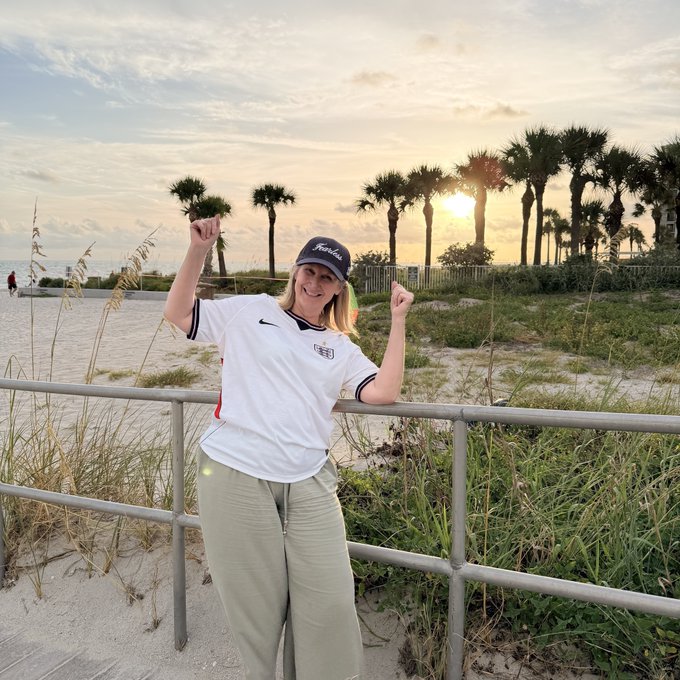 A smiling woman wearing an England national football team jersey and a Titleist cap raises her arms in celebration on a beach boardwalk at sunset, with palm trees visible in the background.