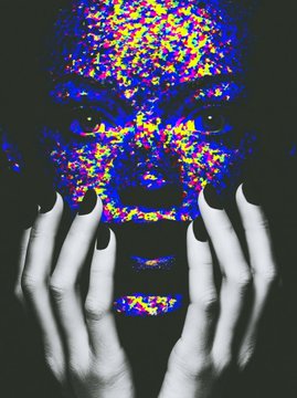 A person's face is partially obscured by their black-manicured hands, with their skin covered in a vibrant, colorful pixelated pattern.