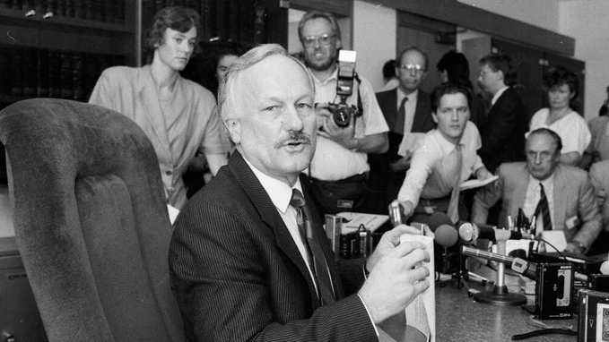 A black and white photograph of a distinguished older man with a mustache sitting at a table with microphones, surrounded by reporters and a photographer in what appears to be a press conference.